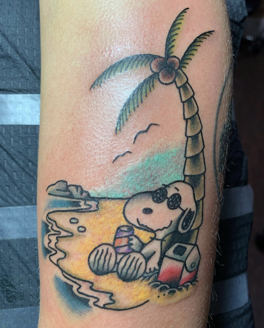 Snoopy smiles while holding a can and sitting against a palm tree on a beach.
